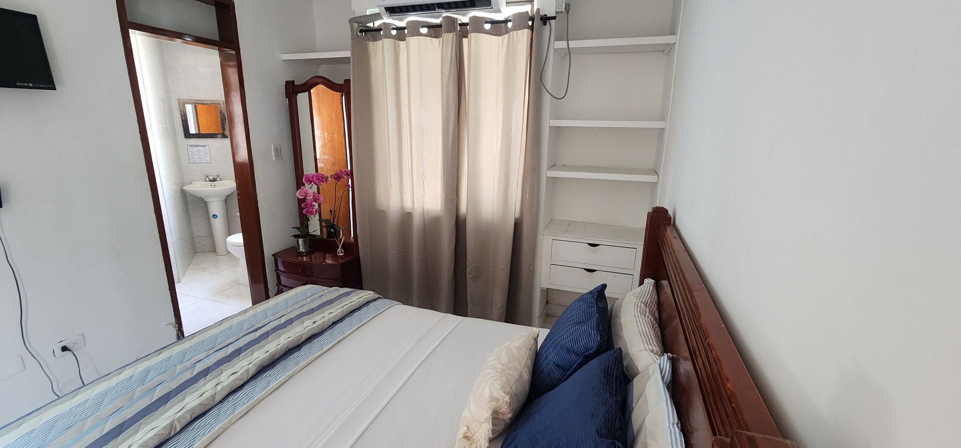 Un dormitorio con cama de matrimonio, ropa de cama azul y blanca, cortinas beige, estanterías empotradas y vistas al baño.