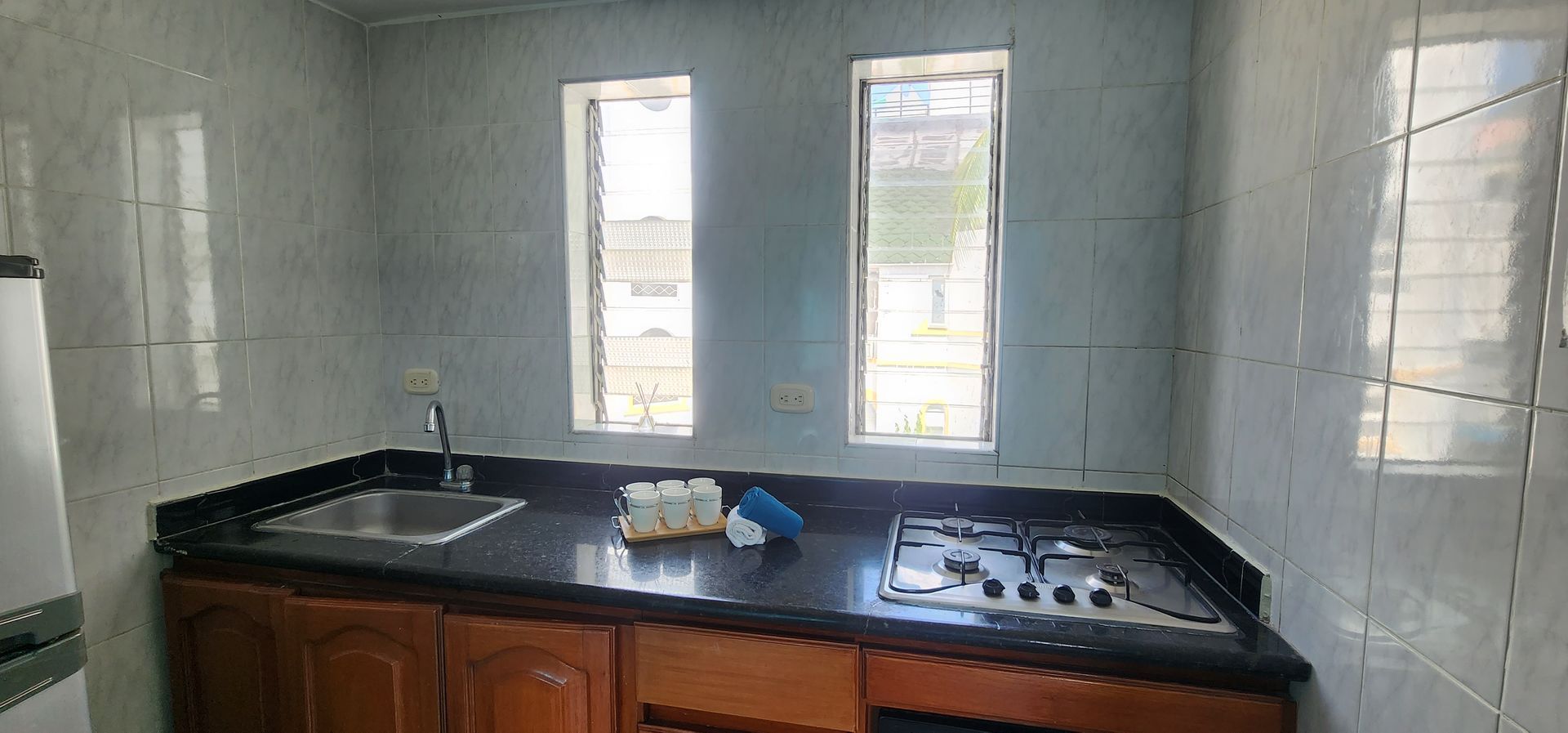 Una encimera de cocina con fregadero de acero inoxidable, superficie de granito negro, placa de gas y dos ventanas en paredes alicatadas.