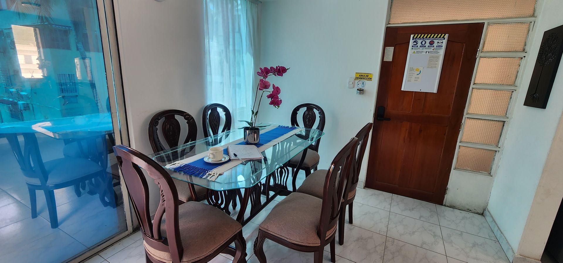 Una mesa de comedor de cristal con cuatro sillas marrones en una habitación muy iluminada, con una pequeña orquídea rosa y una puerta de madera.