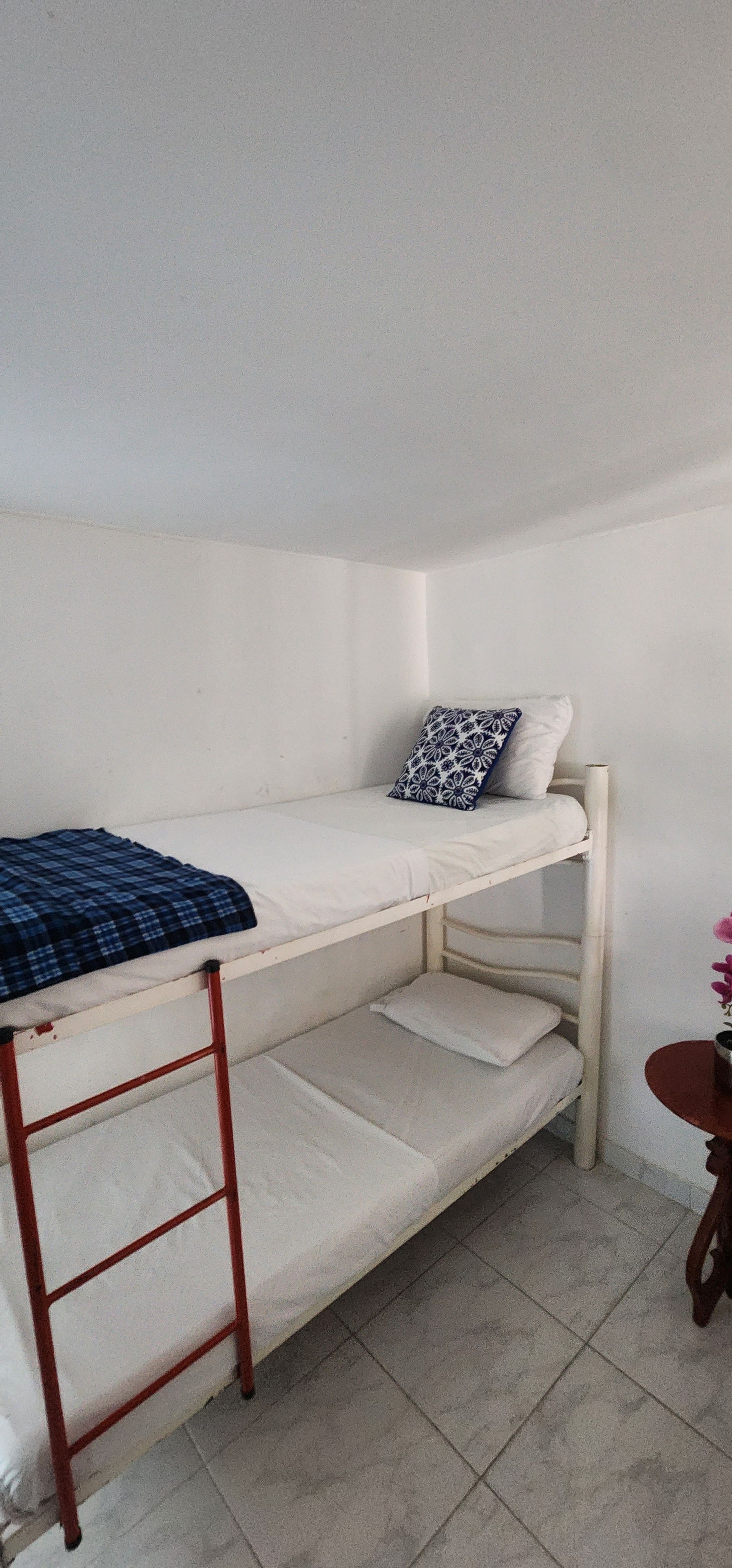 Una litera de metal blanco con almohadas azules estampadas y una manta azul marino en una habitación pequeña con suelo de baldosas de color claro.