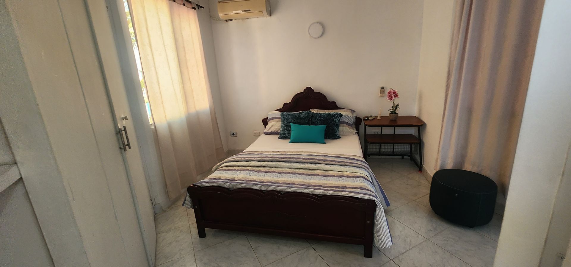 Un dormitorio con una cama de madera oscura, una colcha blanca, almohadas color turquesa, una mesita de noche pequeña con una flor y un puf negro.