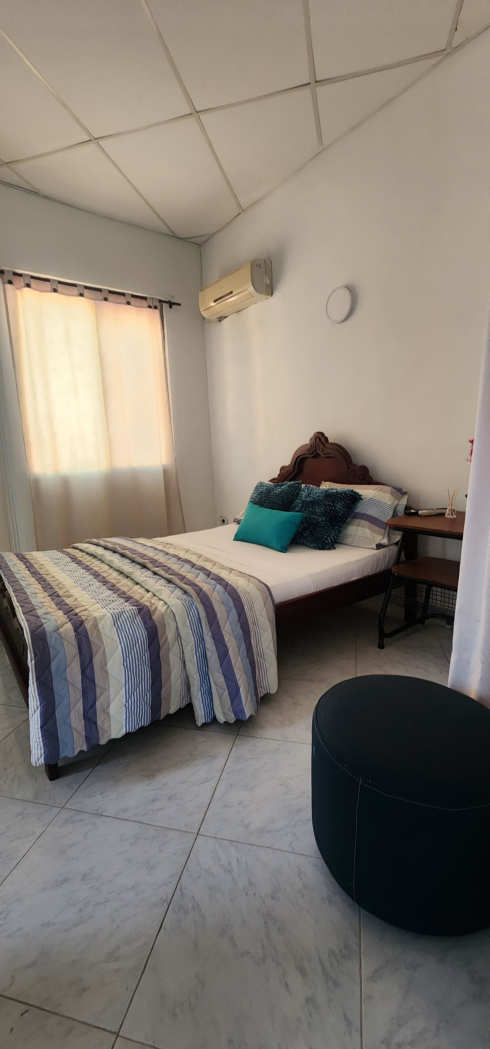 Un dormitorio con una cama cubierta con una manta de rayas azules, una pequeña mesita de noche y un puf negro sobre un suelo de baldosas.