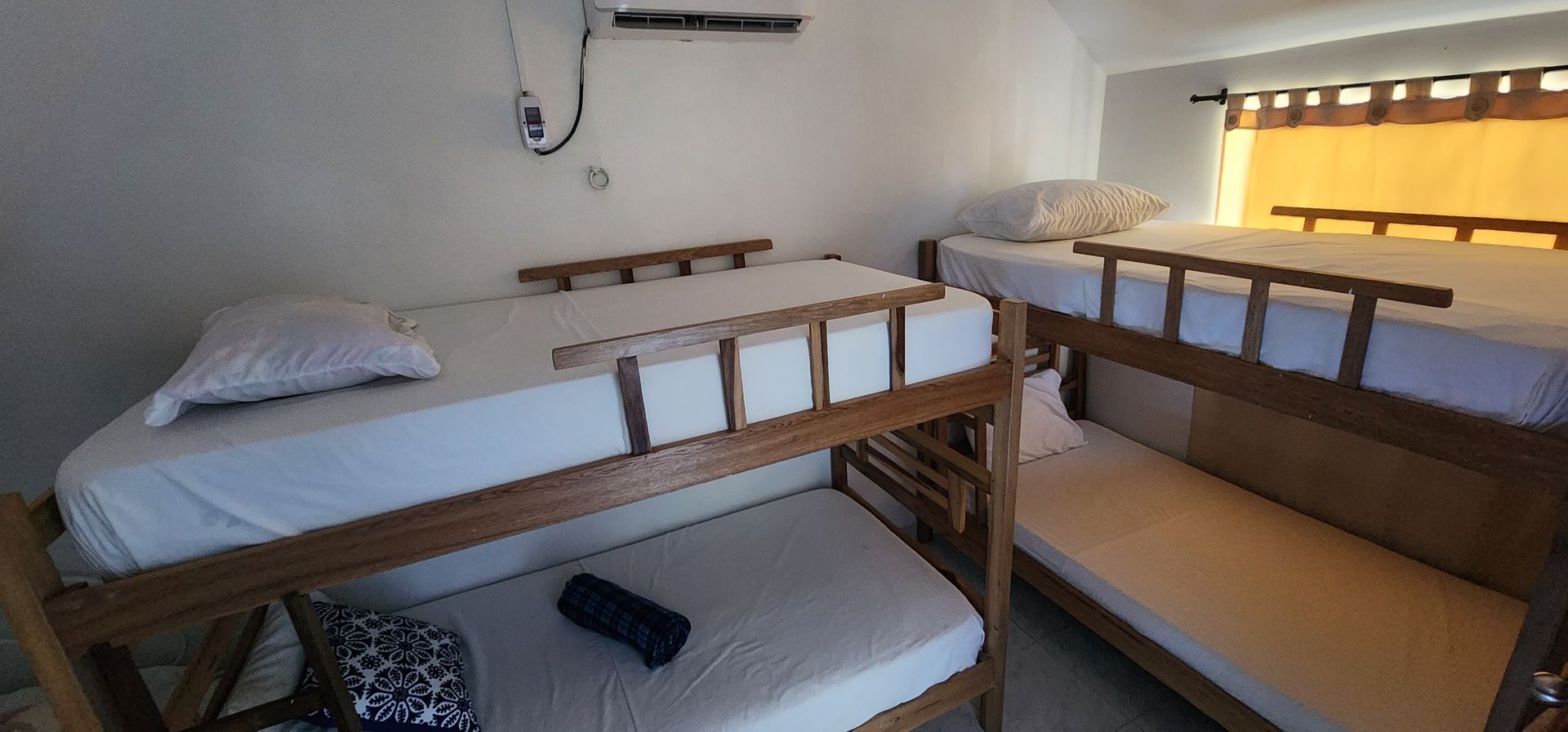 Una pequeña habitación con dos literas de madera con sábanas blancas, una de ellas con una almohada azul oscuro y un cojín estampado.