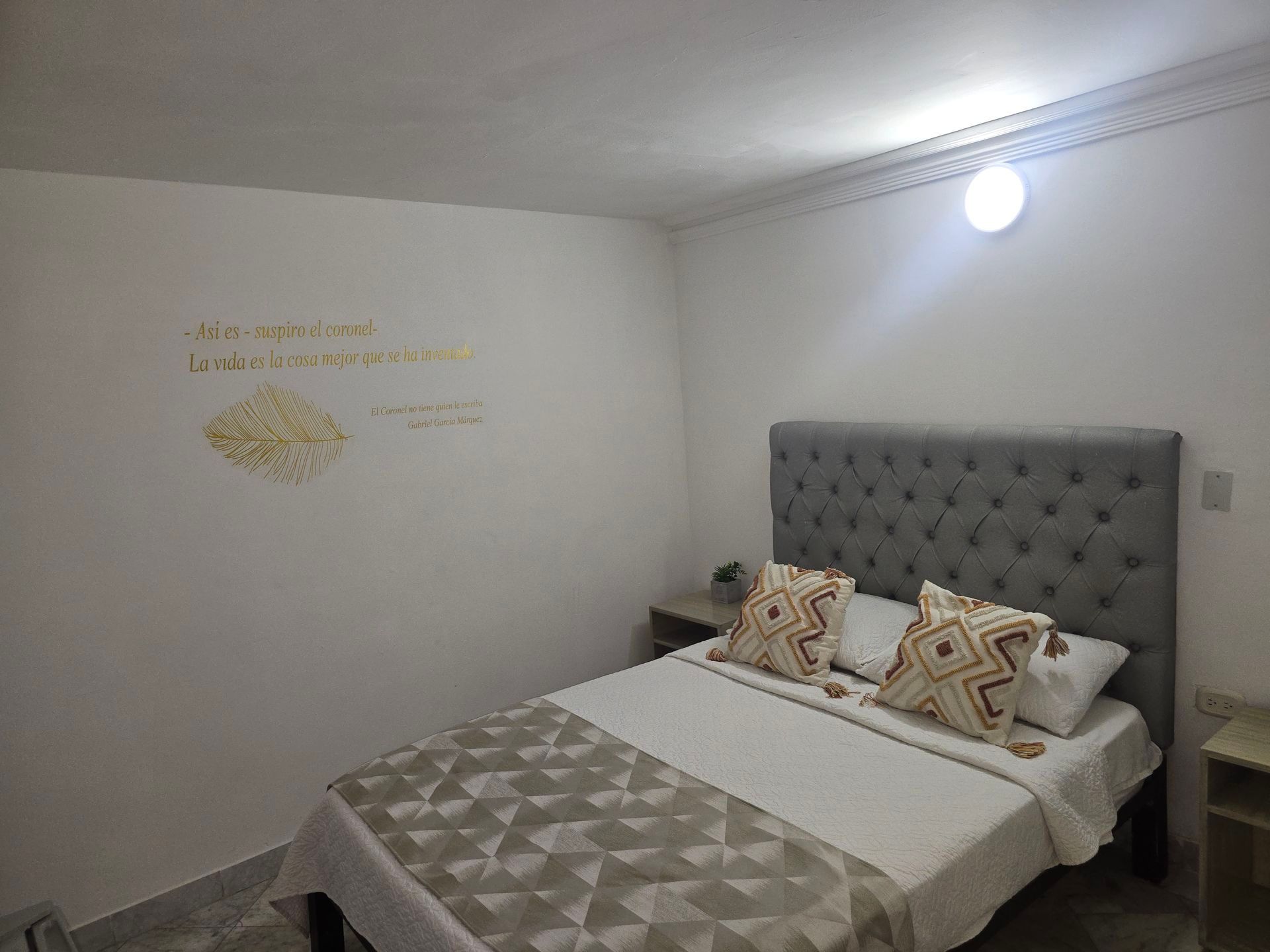 Un dormitorio acogedor con cabecero gris acolchado, ropa de cama blanca con estampado geométrico y texto dorado en la pared.
