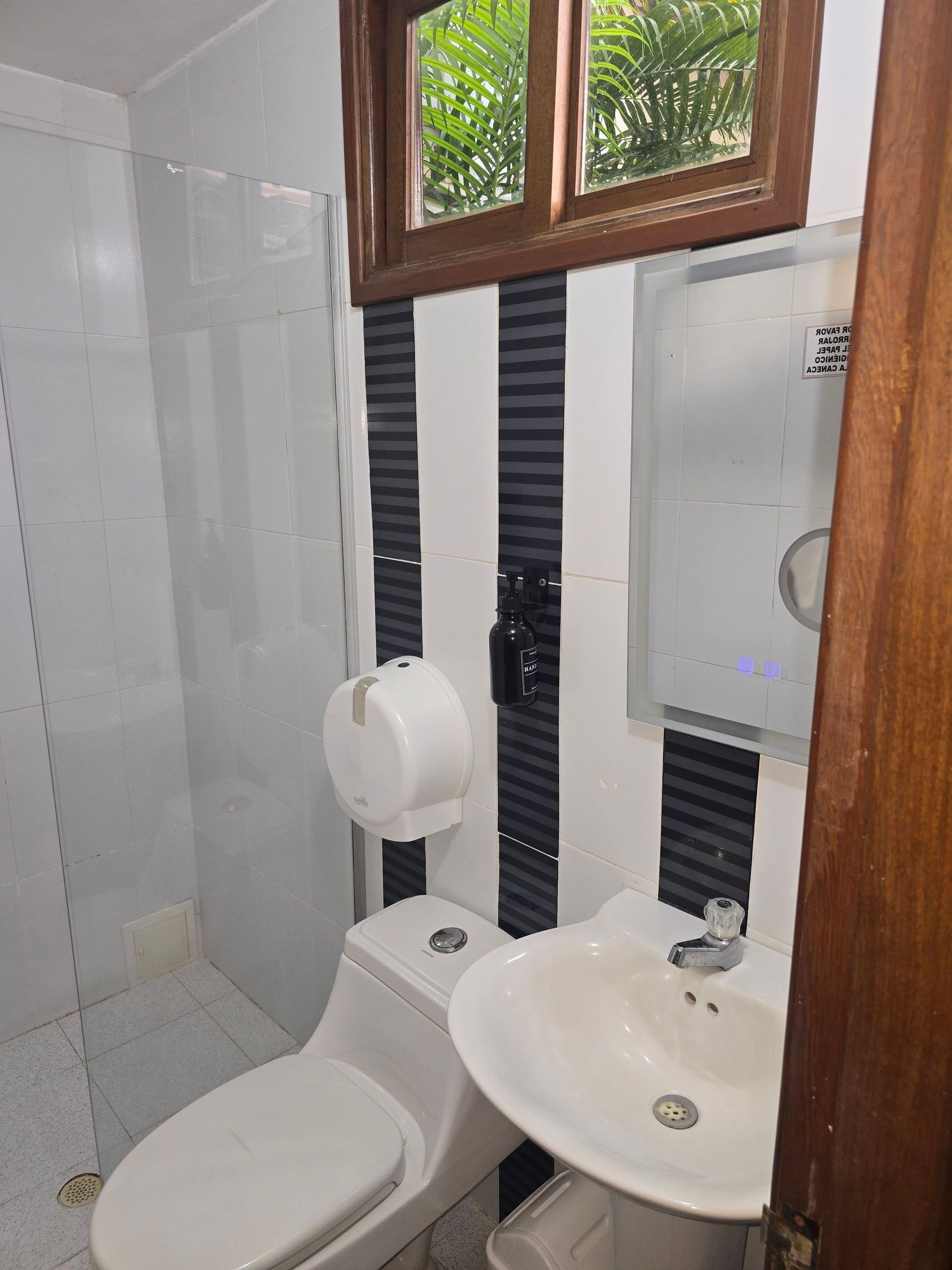 Un baño con azulejos a rayas blancas y negras, un lavabo con pedestal, un inodoro, un dispensador de papel montado en la pared y una ducha.