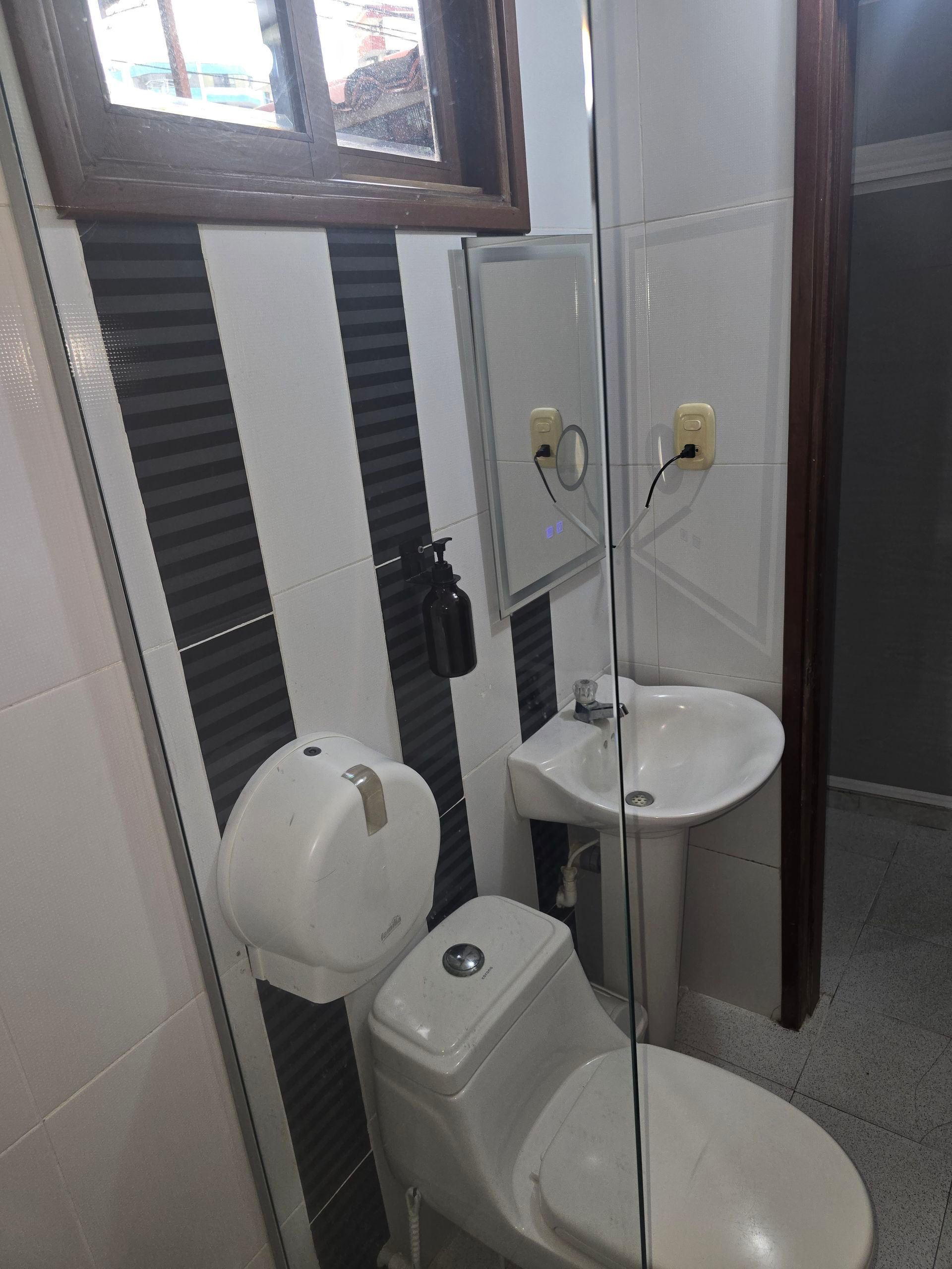 Un baño con inodoro, lavabo con pedestal, dispensador de papel higiénico y una pared a rayas debajo de una ventana.