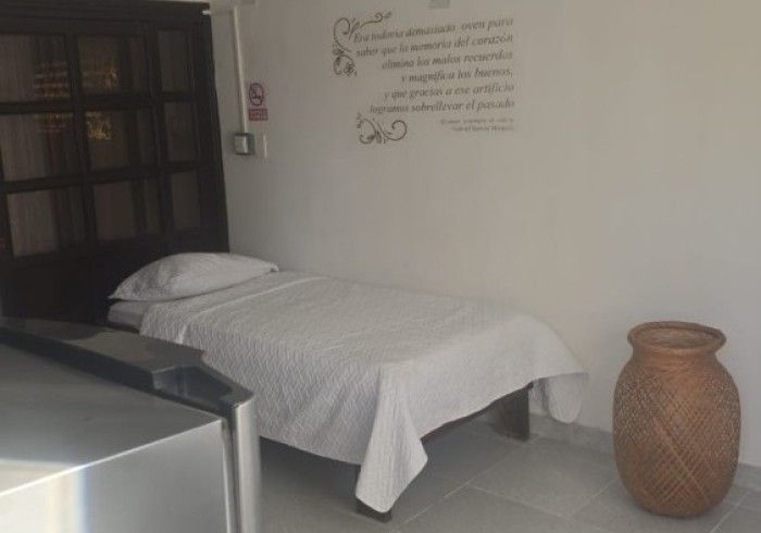 Un dormitorio minimalista con una cama individual con sábanas blancas, un jarrón de suelo con textura y una cita en la pared.