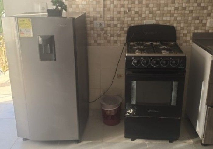 Una zona de cocina con un frigorífico plateado a la izquierda, una estufa negra a la derecha y un pequeño cubo de basura rojo en medio.