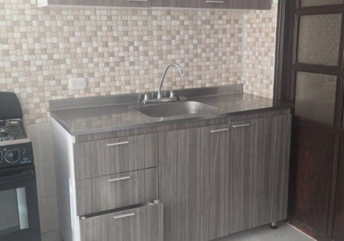 Una encimera de cocina con un mueble de madera gris, un fregadero de acero inoxidable y un protector contra salpicaduras de azulejos junto a una estufa de gas.