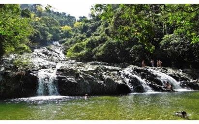 Una cascada cae sobre rocas oscuras hasta una poza de agua verde cristalina, rodeada de un denso bosque y con gente cerca.