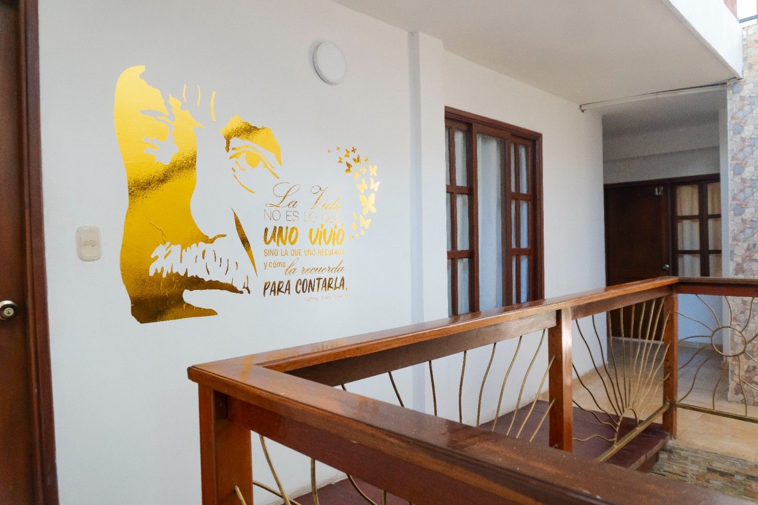 Un pasillo con una silueta dorada estampada en la pared blanca, junto a una barandilla de balcón de madera y marcos de ventanas.
