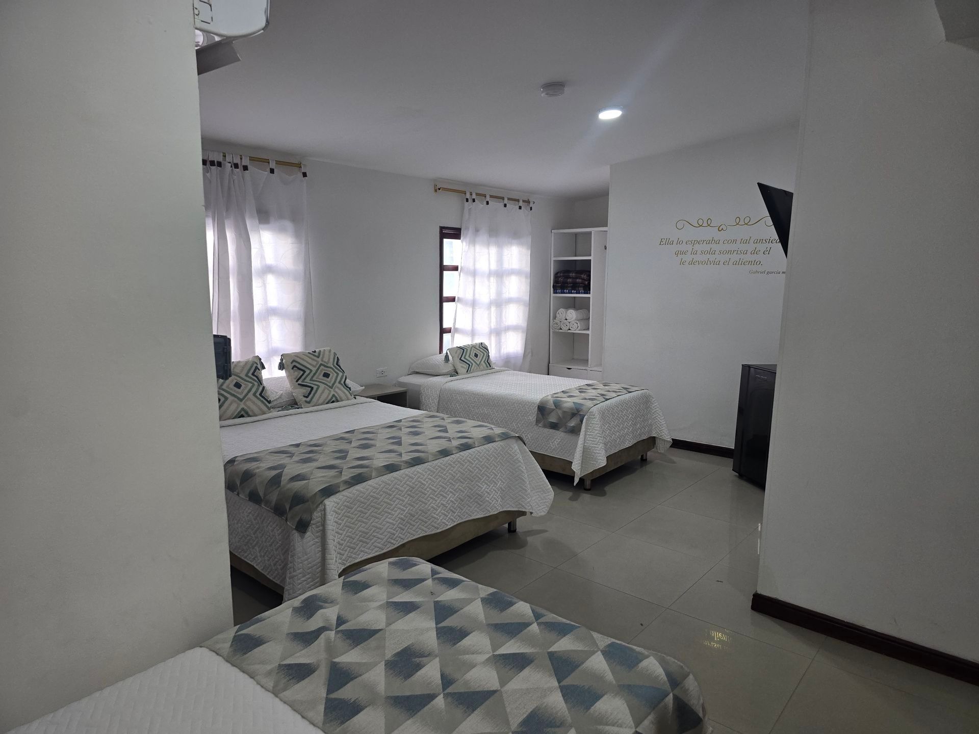 Una habitación de hotel con tres camas, ropa de cama estampada en gris y blanco, paredes blancas y cortinas transparentes.