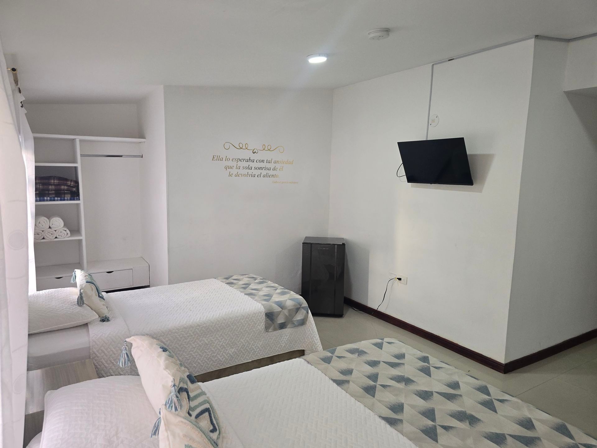 Una habitación de hotel con dos camas blancas, un armario abierto, un televisor montado en la pared y una cita decorativa en la pared.