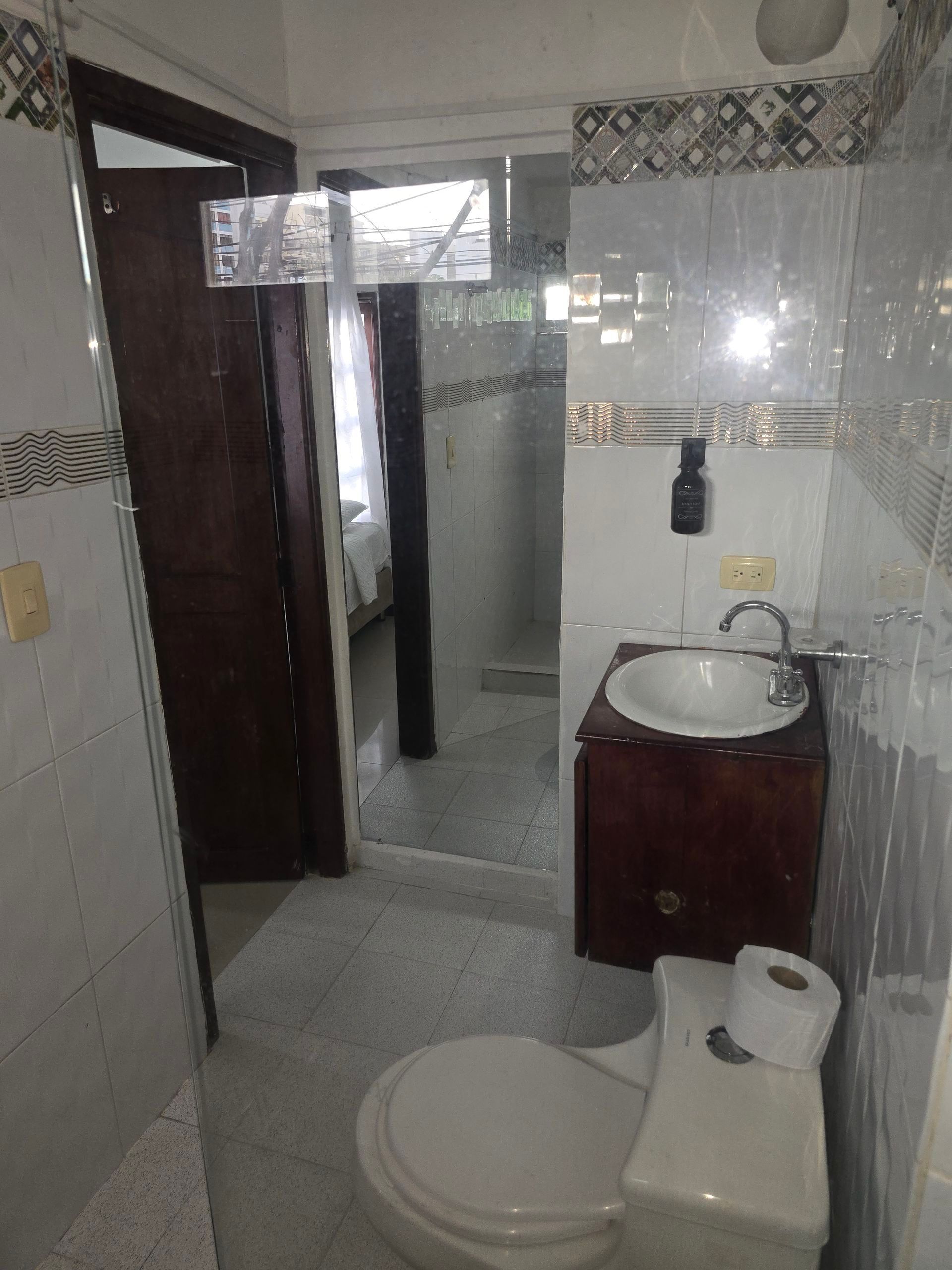 Un baño con inodoro blanco, tocador de madera con lavabo blanco sobre encimera, paredes alicatadas y una puerta abierta que da a la ducha.