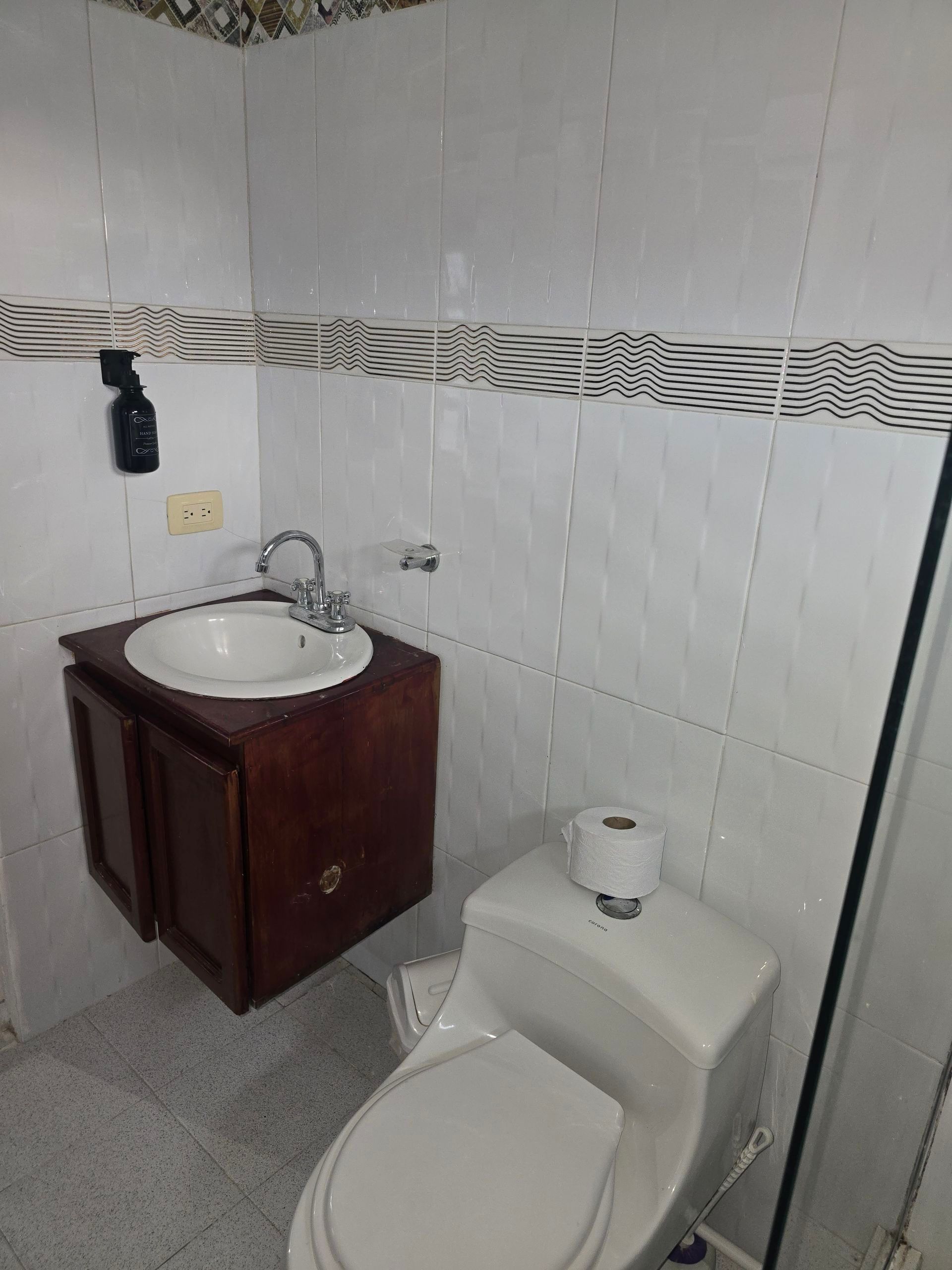 Un baño pequeño con paredes alicatadas en blanco, un tocador de madera oscura, un lavabo redondo y un inodoro blanco.