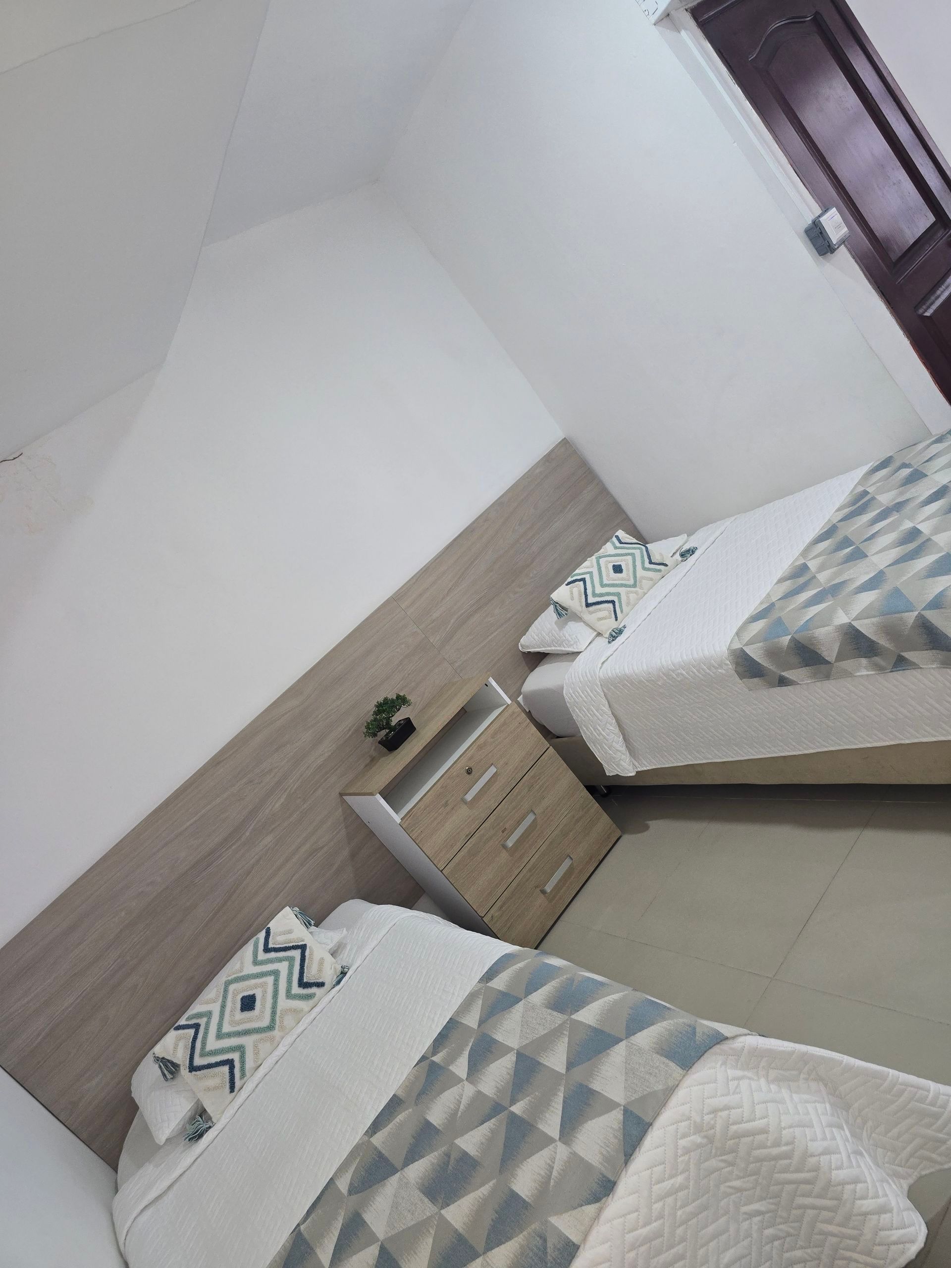 Una habitación de hotel con dos camas individuales cubiertas con mantas estampadas grises y blancas, una pequeña mesita de noche de madera y una puerta oscura.