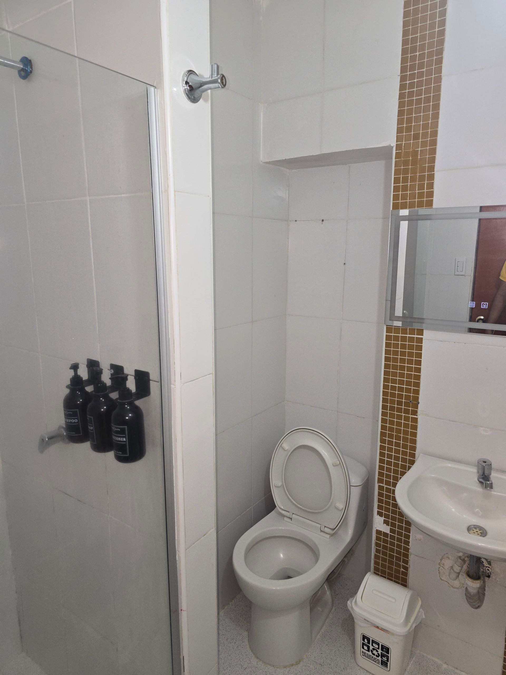 Un baño con azulejos blancos que cuenta con una zona de ducha con botellas instaladas, un inodoro central y un lavabo con azulejos con motivos dorados.