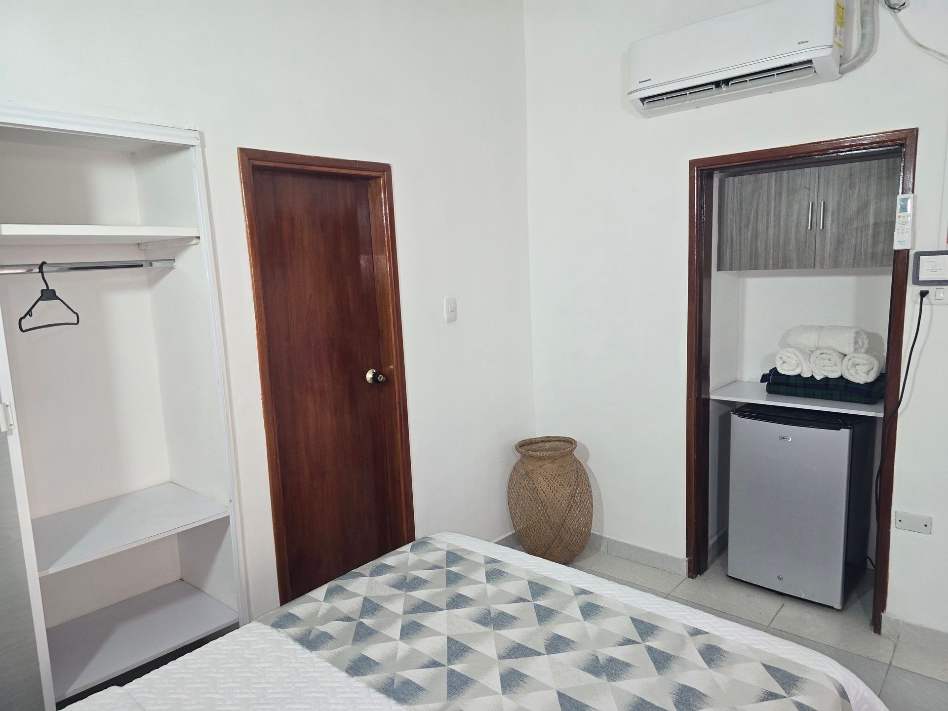 Una habitación de hotel con una colcha estampada, un armario abierto, una puerta de madera, una cesta de mimbre y una mininevera debajo de un estante.