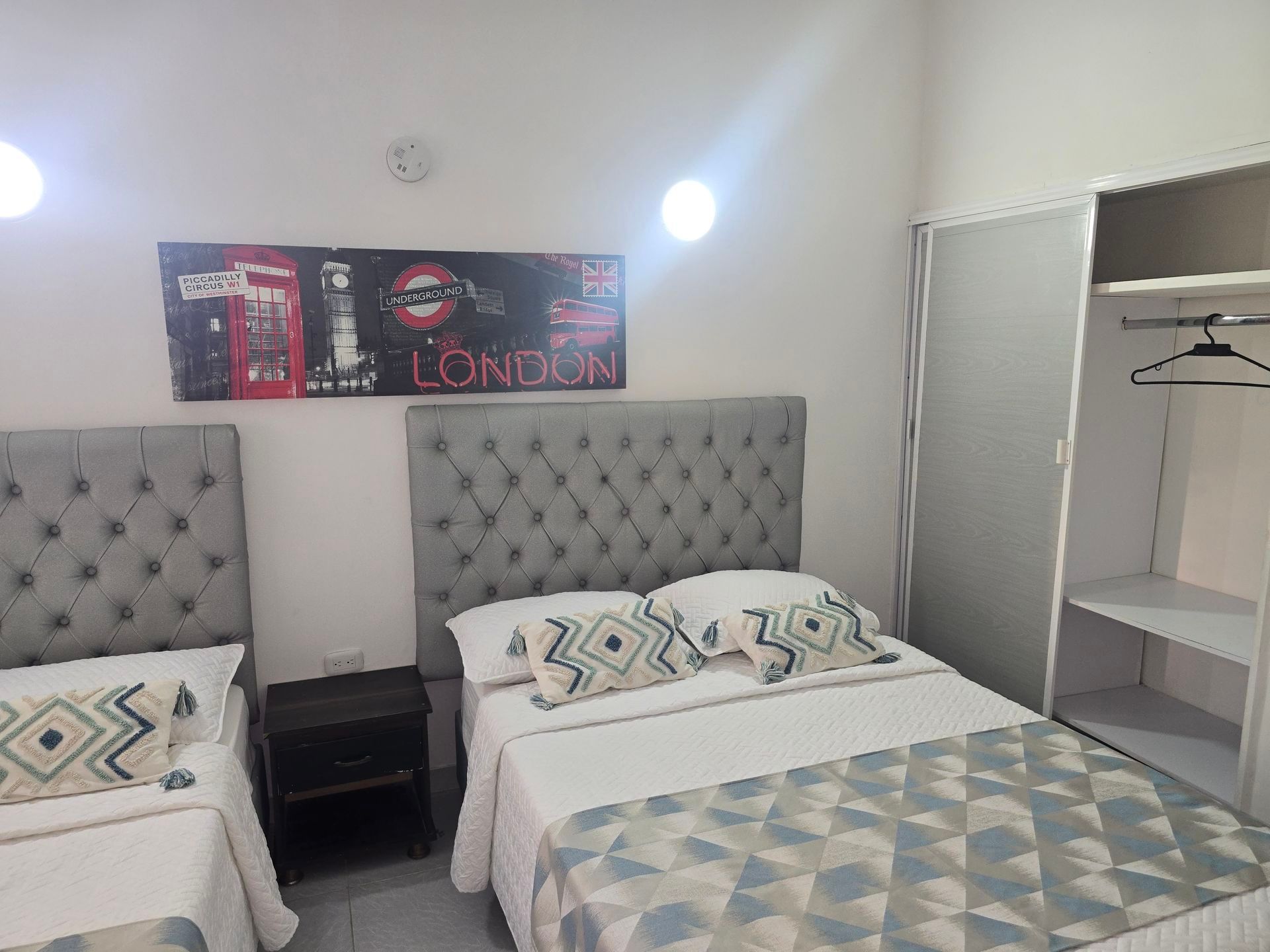 Un dormitorio con dos camas que cuentan con cabeceros acolchados grises, cojines estampados, una mesita de noche y un cuadro con motivos londinenses en la pared.