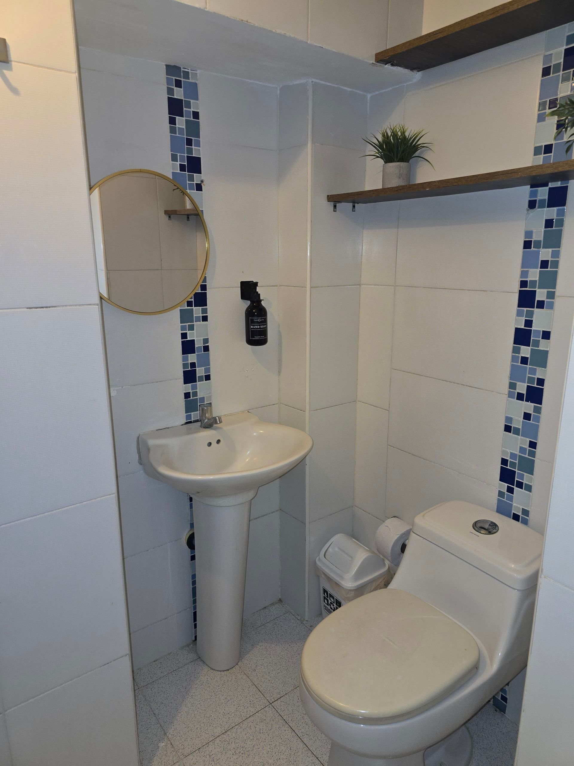 Un pequeño baño con lavabo de pedestal blanco, inodoro y dos estantes de pared con detalles decorativos de azulejos azules.