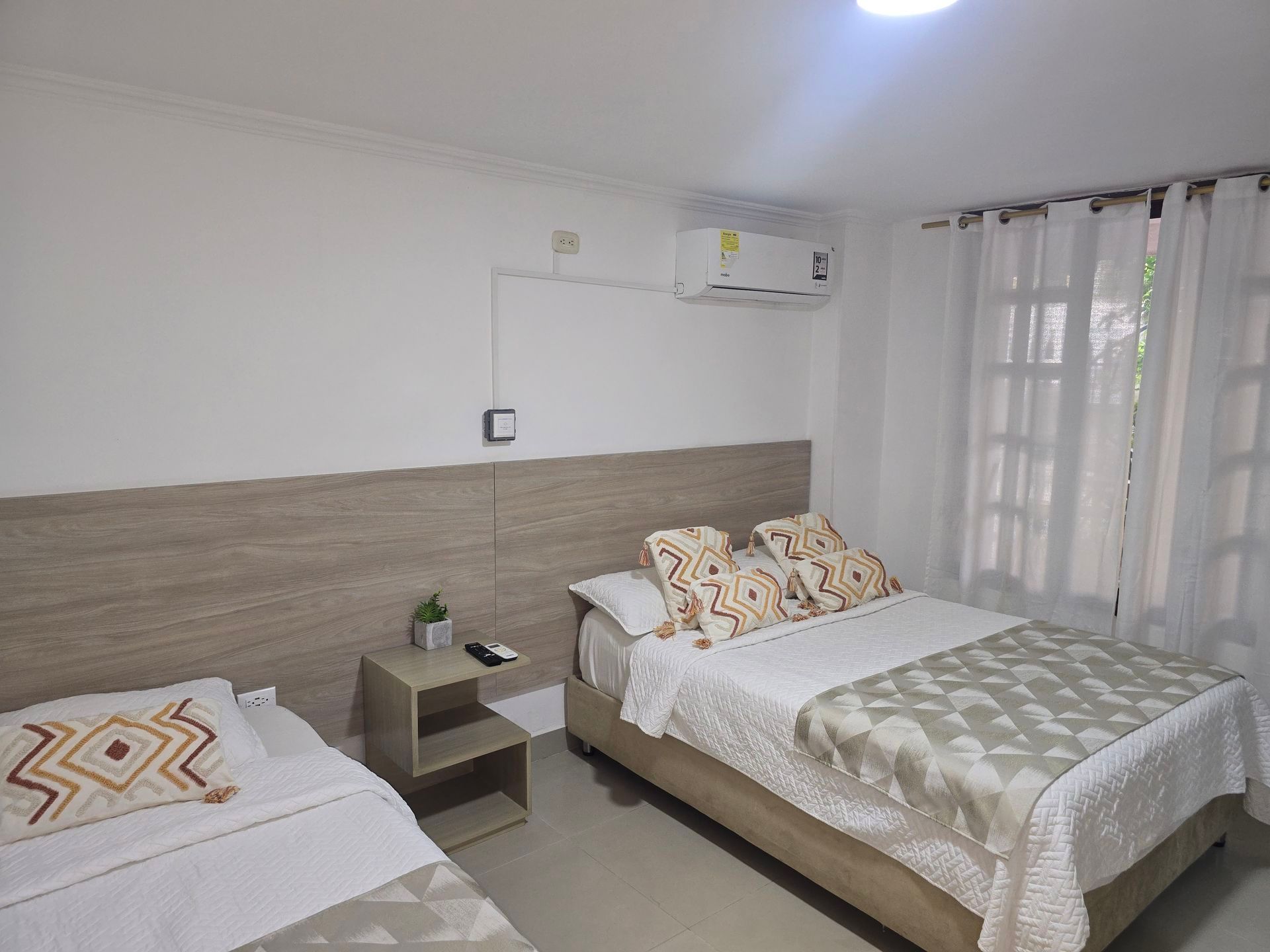 Una habitación de hotel con dos camas, un cabecero de madera, una mesita de noche pequeña y cortinas blancas que cubren una ventana.