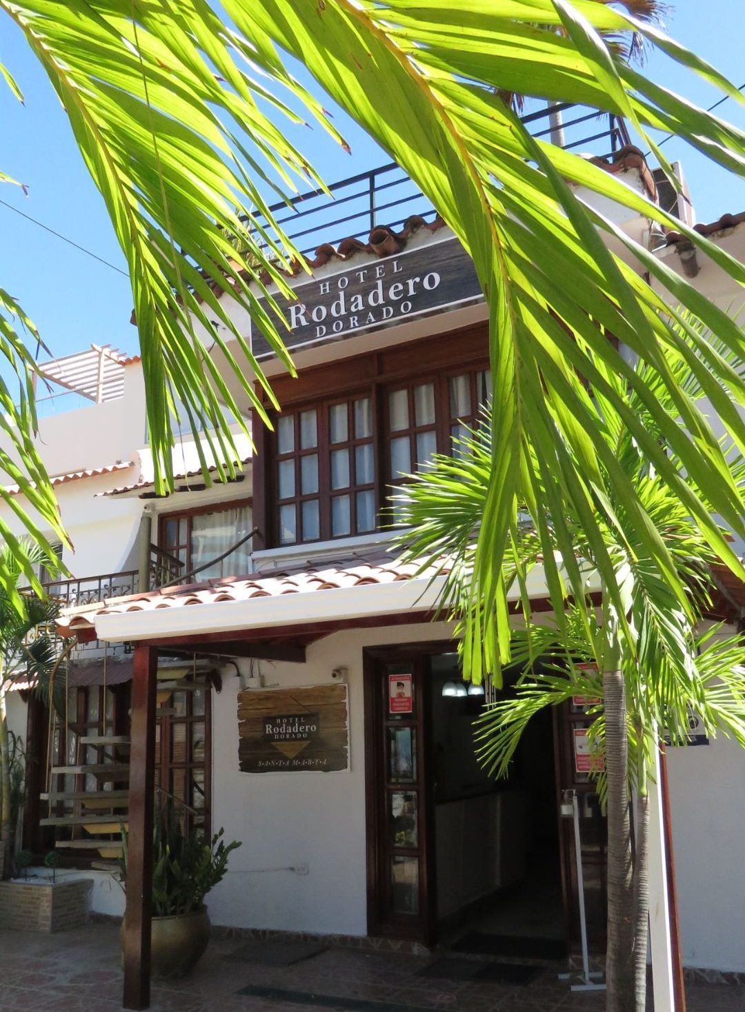 Exterior del Hotel Rodadero con paredes blancas, detalles de madera y hojas de palmera 