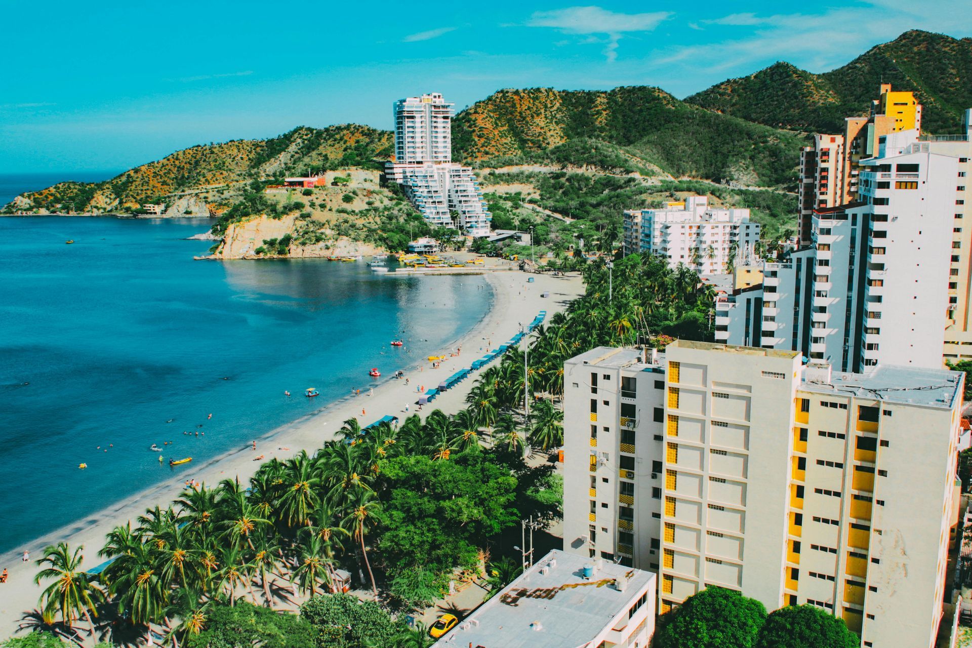 Horizonte urbano costero junto a una bahía de aguas turquesas y una playa de arena, con palmeras y edificios en la ladera.