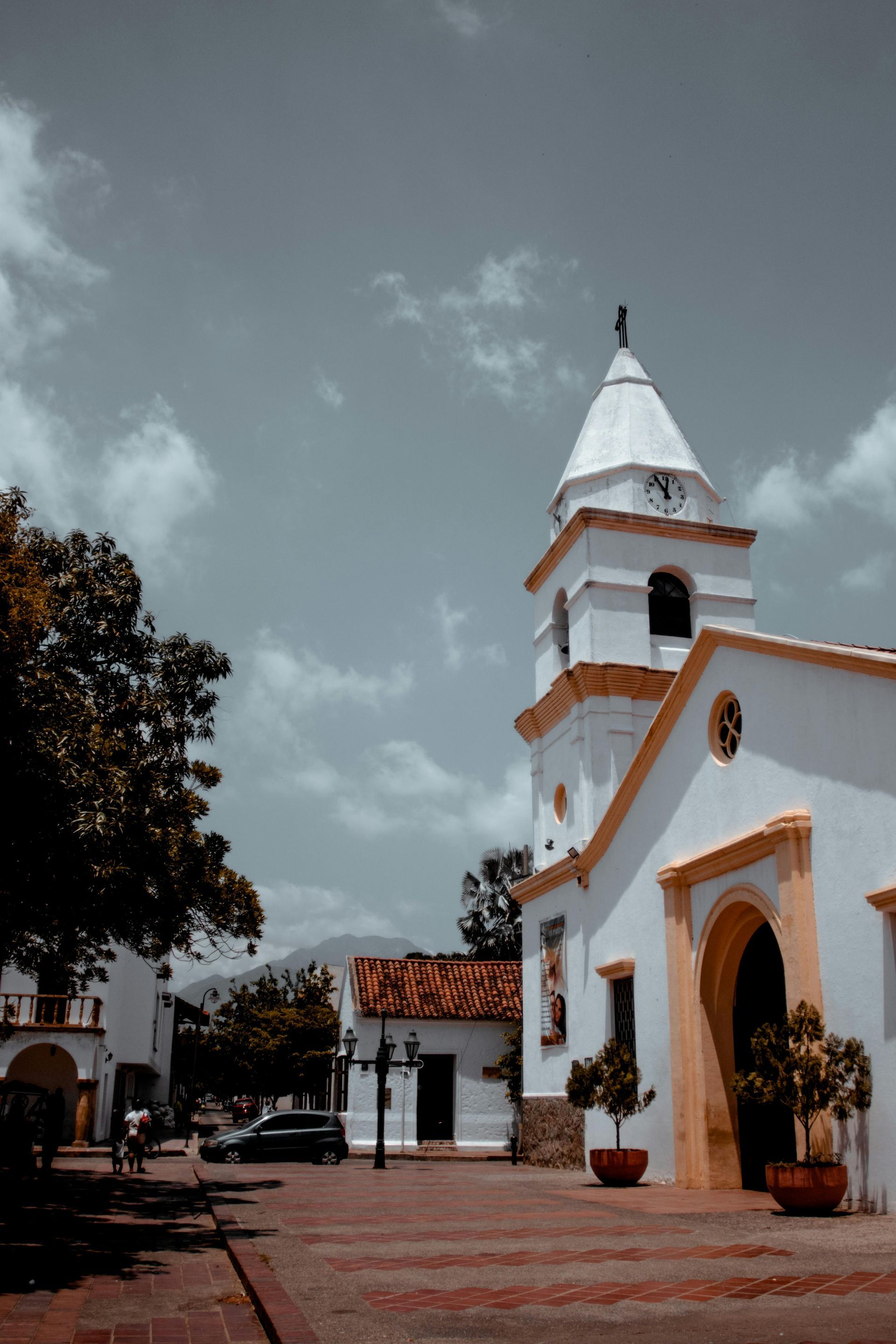Una iglesia colonial blanca con campanario y detalles amarillos se alza en una plaza pavimentada bajo un cielo nublado.