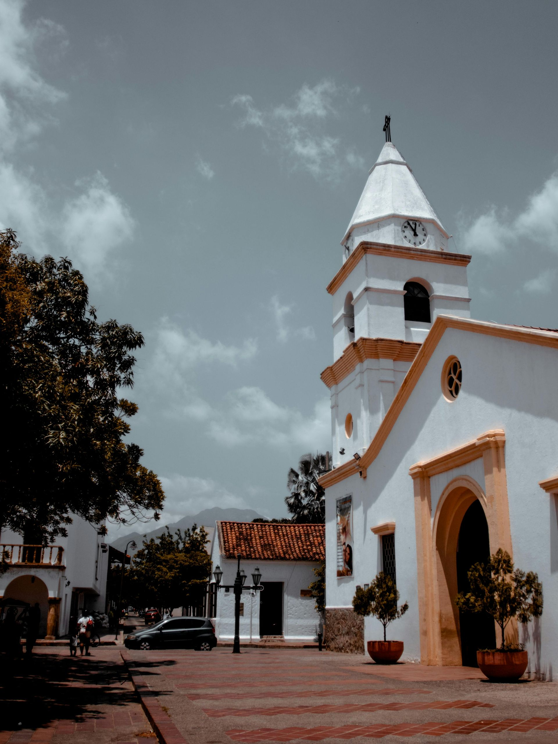 Iglesia colonial blanca con campanario, detalles en naranja y plantas en macetas en una plaza empedrada bajo un cielo nublado.