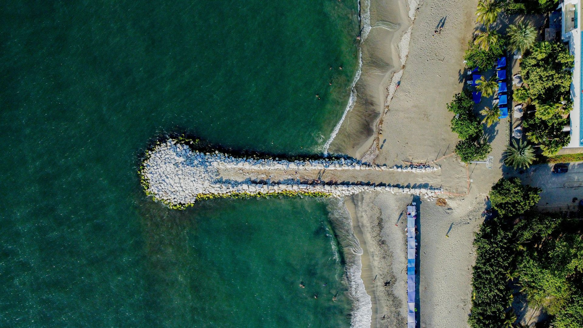 Vista aérea de un embarcadero de piedra que se adentra en aguas azul oscuro desde una playa de arena bordeada de árboles verdes.