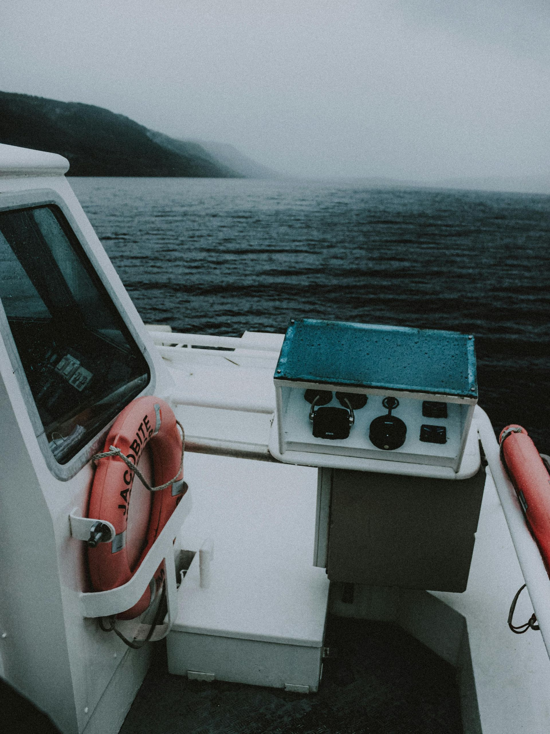 La cubierta de un barco con vistas a colinas lejanas y brumosas sobre aguas oscuras, donde se encuentra una consola de control y un salvavidas.