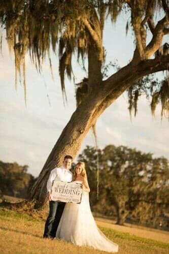 A bride and groom are standing under a tree holding a sign.