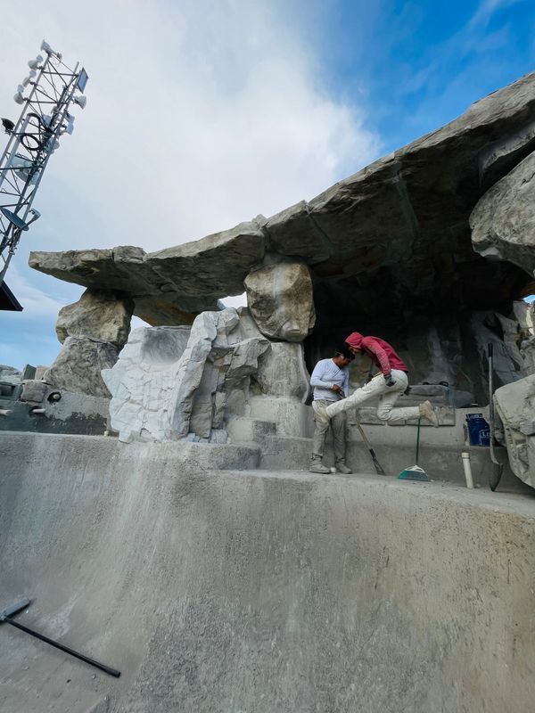 Two people work on a concrete structure with stone-like details. Cloudy sky background.