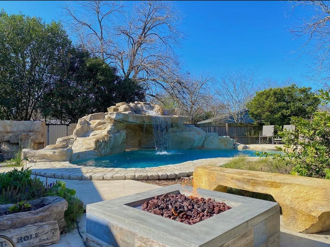 Backyard pool with rock waterfall, fire pit, and seating. Blue water, clear sky.