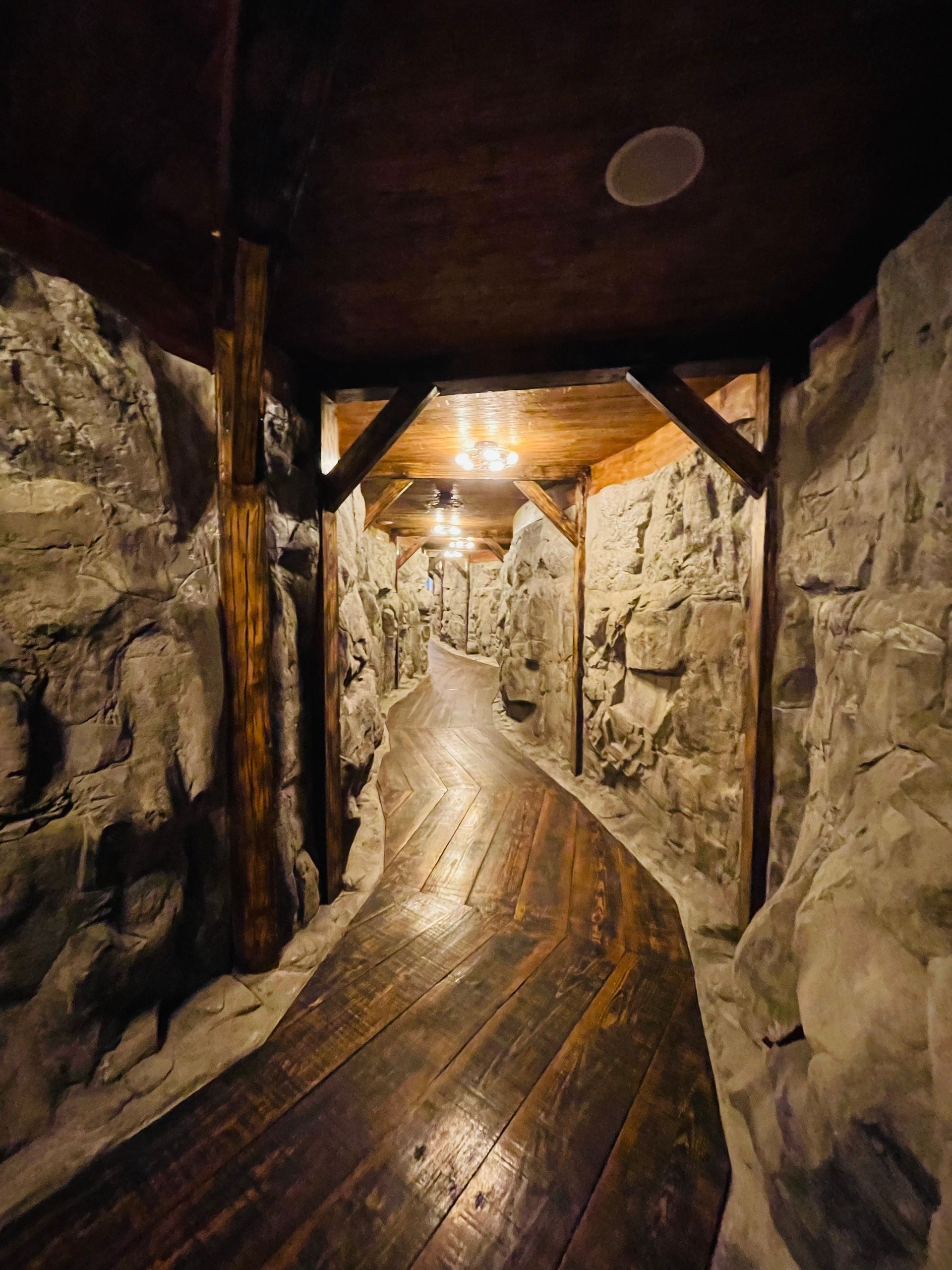 A narrow wooden walkway winds through a stone-walled tunnel, lit by overhead lights.