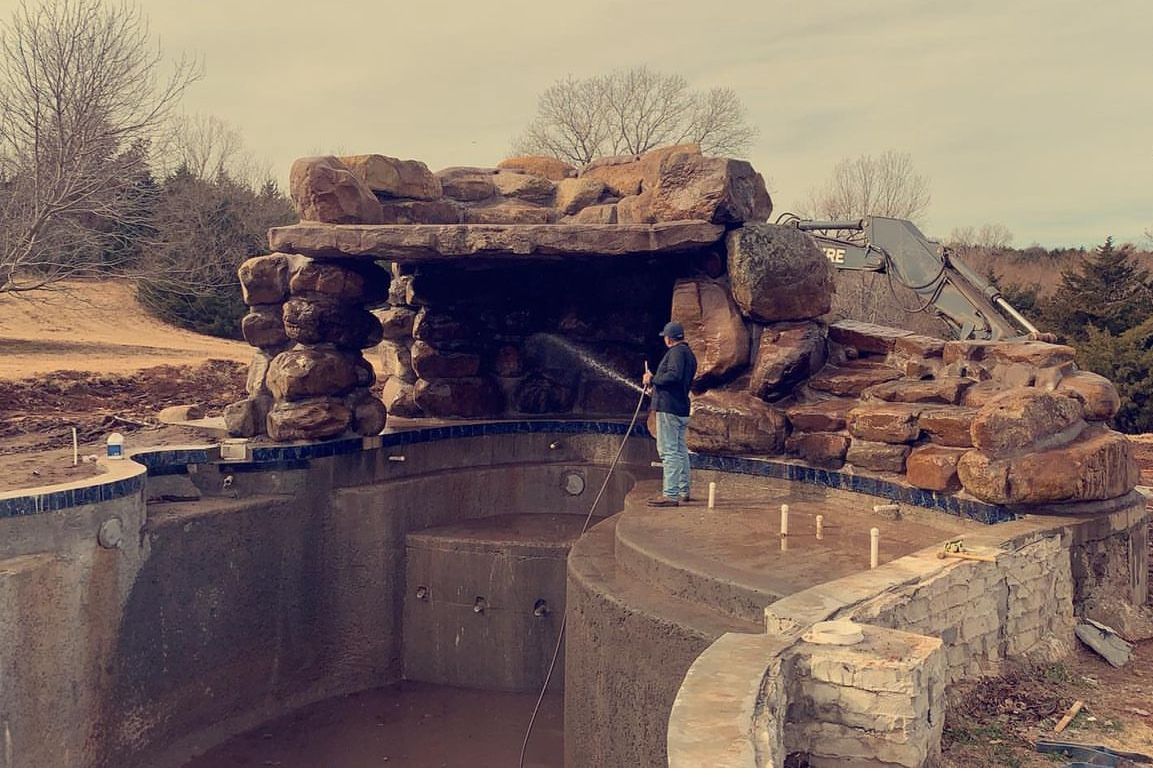 Man spraying a pool with a rock waterfall feature. The pool is empty. Overcast day.