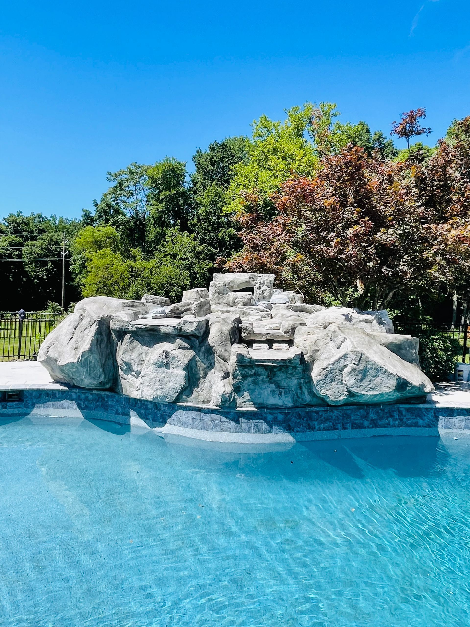 Rock waterfall feature in a blue-tiled pool, against a background of trees under a bright blue sky.
