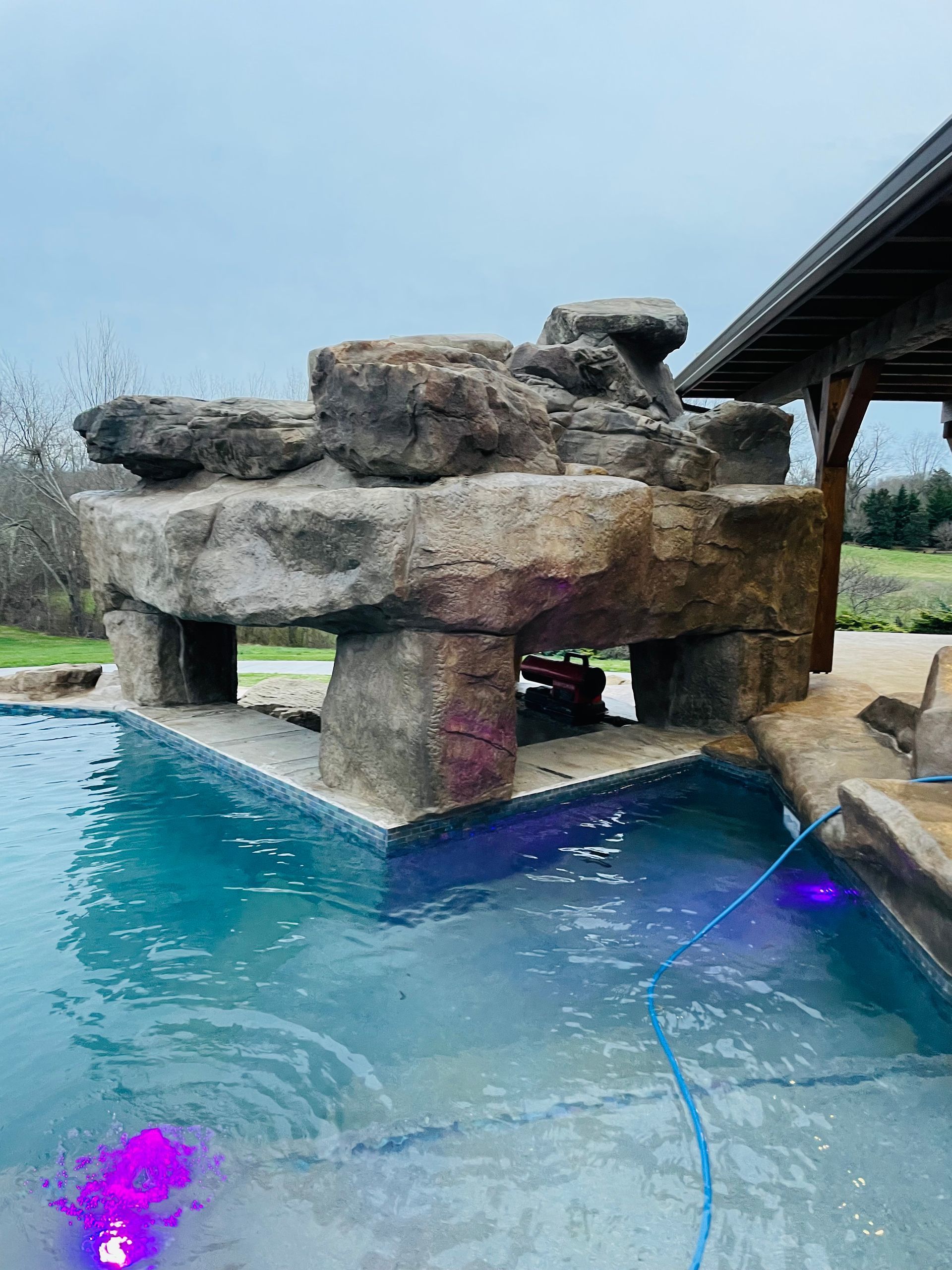 Artificial rock structure over pool water. Blue pool, stone supports, and a covered patio are also visible.