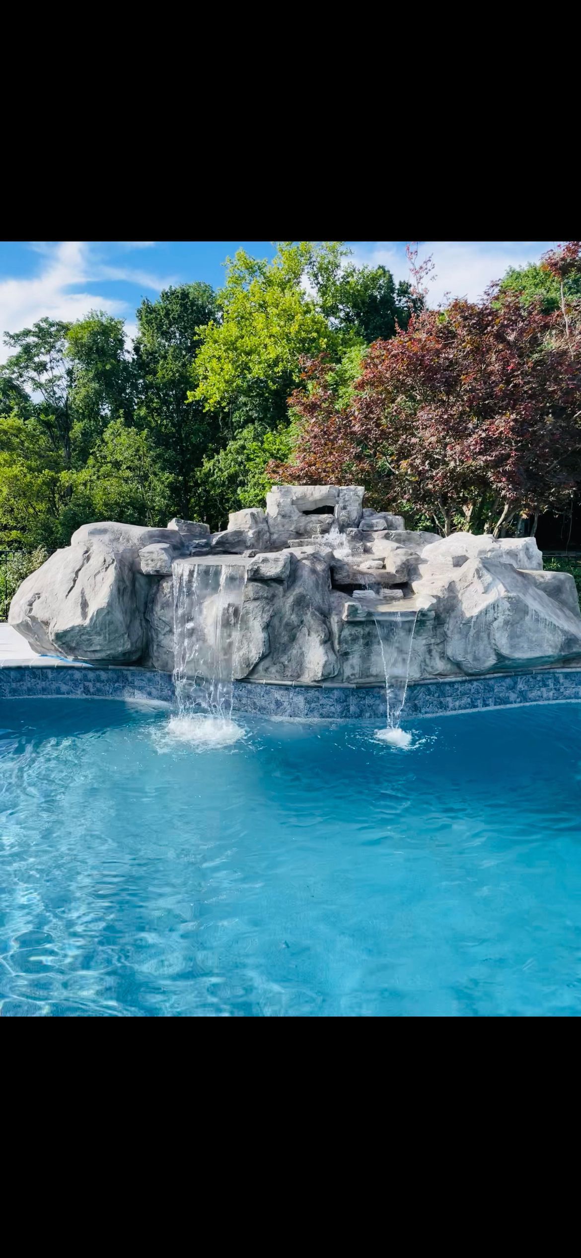 Waterfall feature in a pool, surrounded by trees under a blue sky.