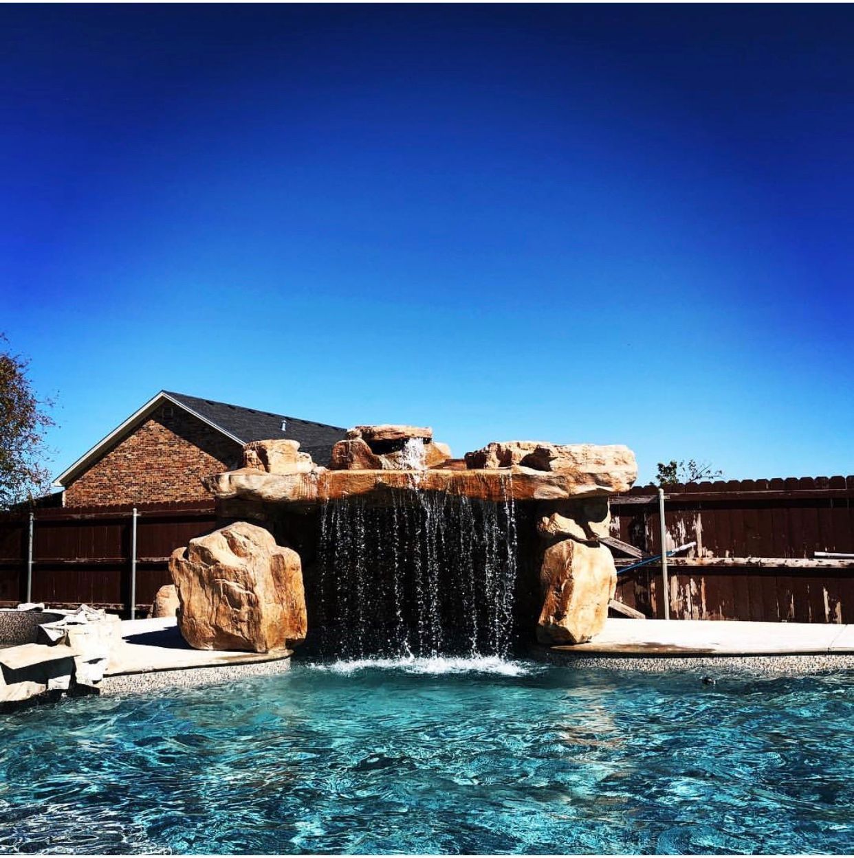 Swimming pool with waterfall feature, brown rocks, blue water, clear sky.