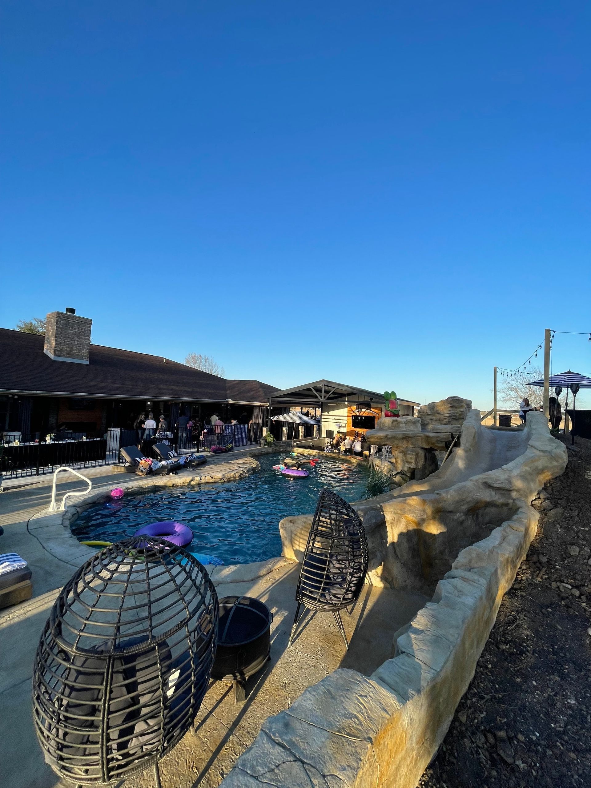 Poolside gathering under a clear blue sky, with a house, bar, and rock features visible.