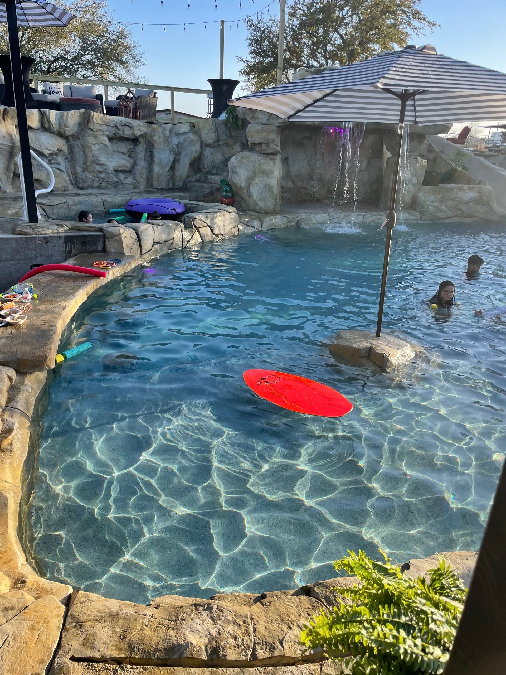 Pool with rock waterfall, red boogie board, and people swimming on a sunny day.