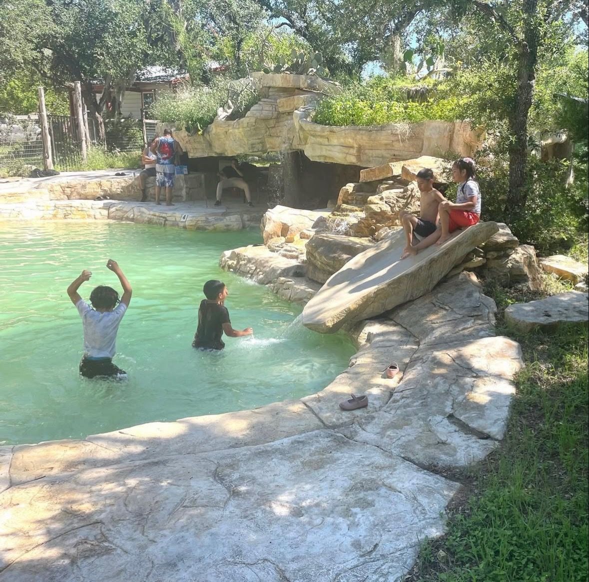 Children playing in a natural-looking pool with stone edging and a small waterfall. Sunny day.