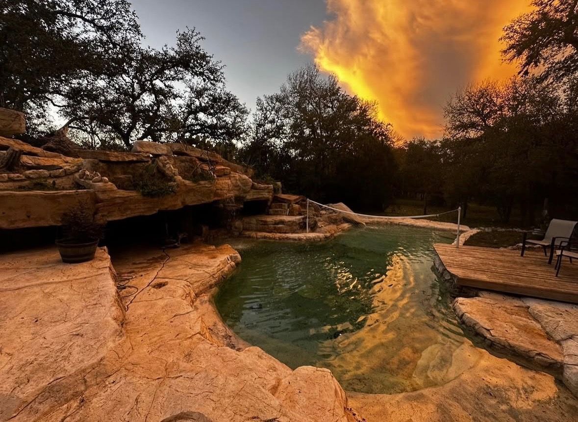 Rock-lined pool with waterfall at dusk, trees surround, fiery orange and yellow sky.