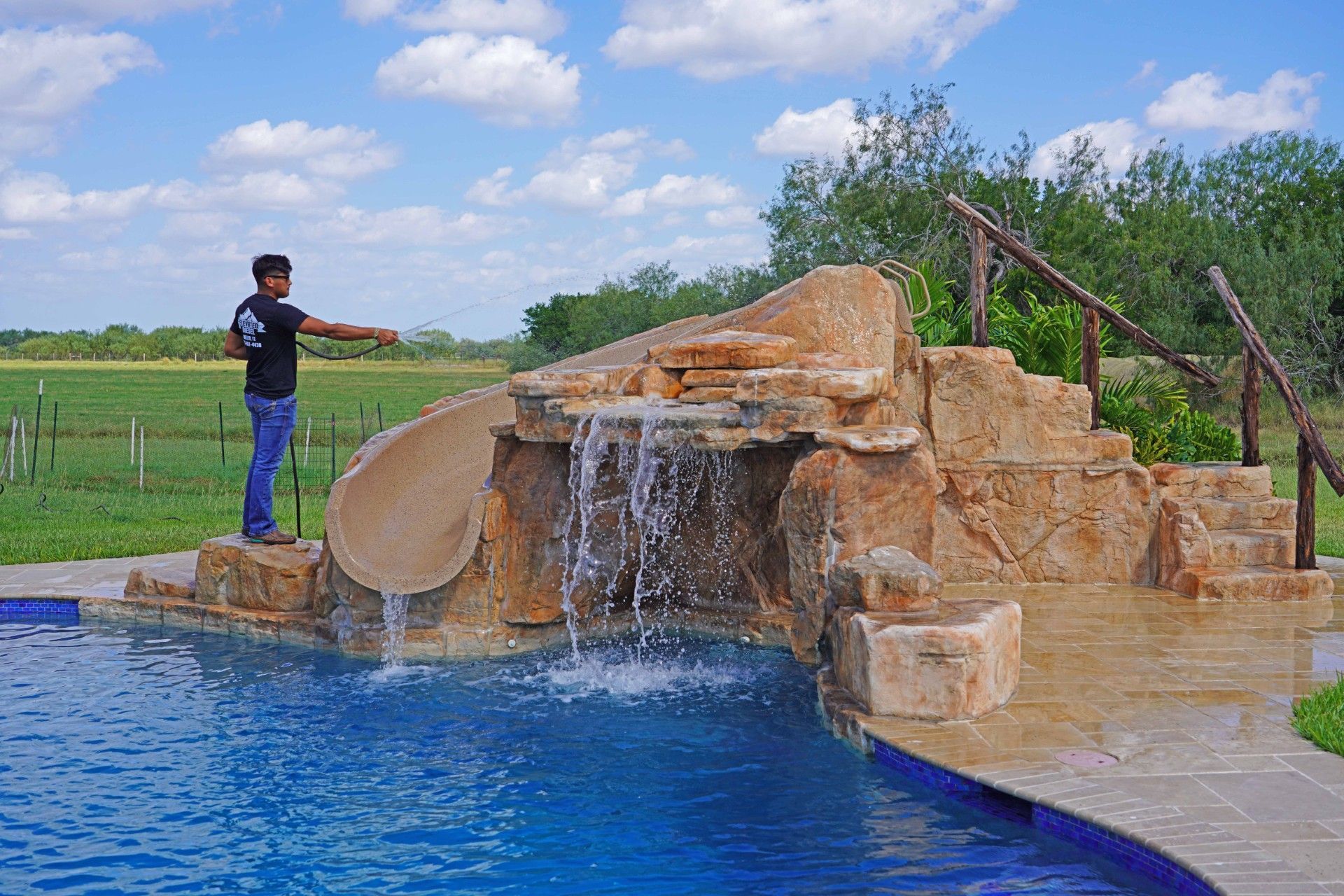 Man standing next to a pool with a water slide and waterfall feature on a sunny day.