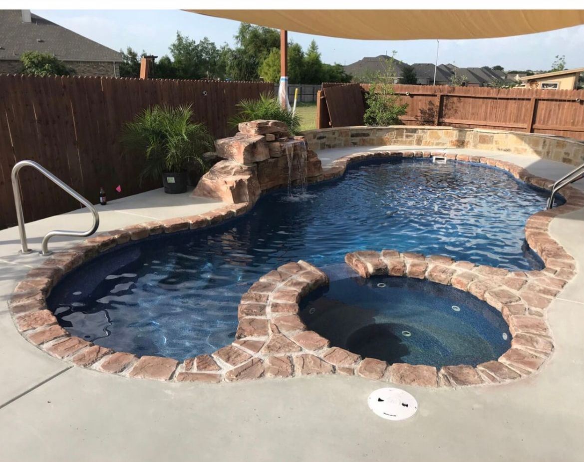 Pool with a waterfall feature, hot tub, and brown brick coping surrounded by a concrete patio.