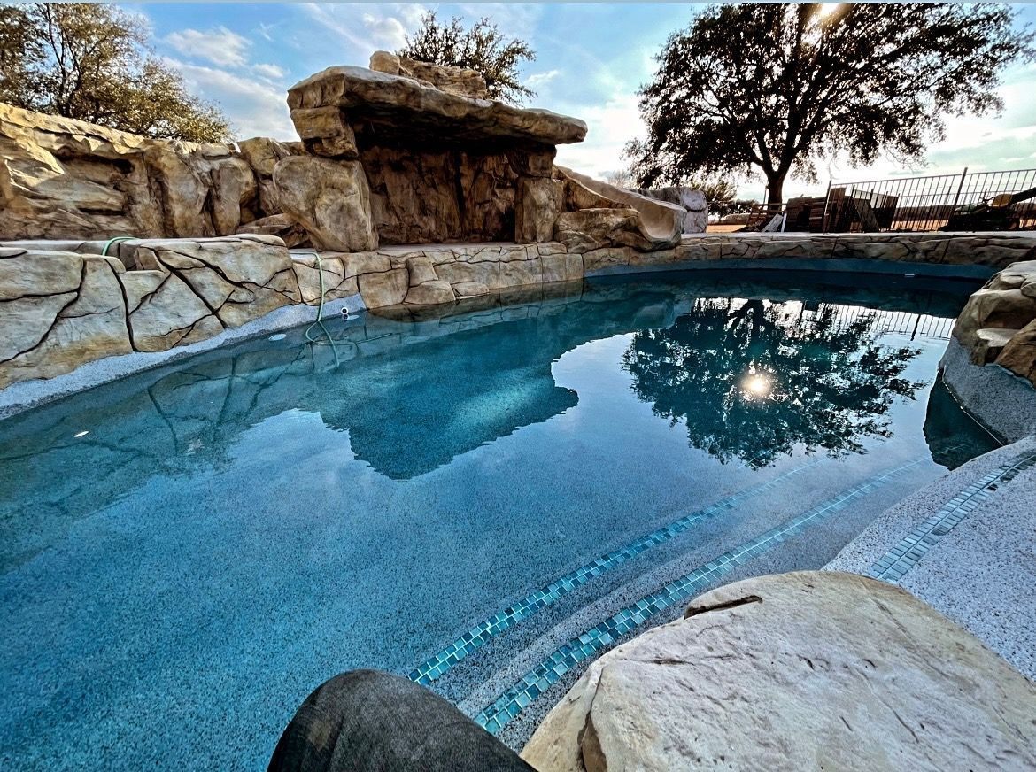 Swimming pool with rock waterfall and surrounding stonework, under a tree in outdoor setting.