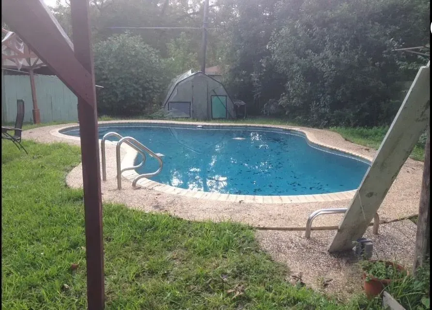 Swimming pool with light blue water and stone deck surrounded by green grass and trees.