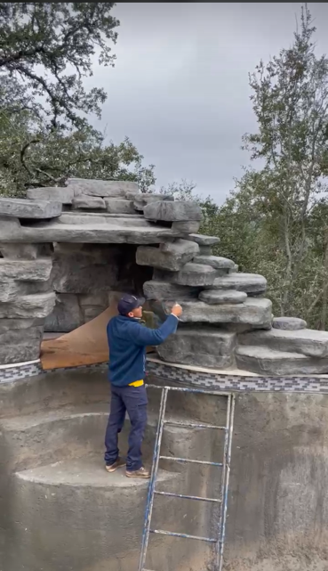 Man on a ladder working on a stone structure near a pool, cloudy day.
