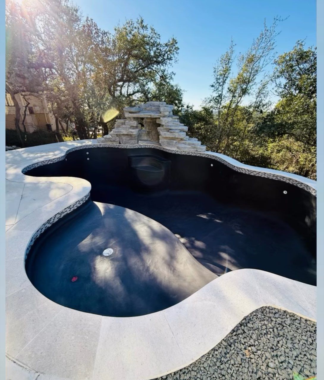 Empty, kidney-shaped pool with a waterfall feature surrounded by concrete and gravel, in a natural setting.